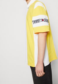 Yellow and white polo shirt with a collar, featuring a contrast stripe and "TOMMY JEANS" logo on the sleeve. Cotton material, relaxed fit.