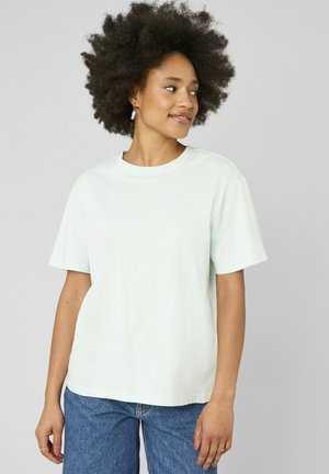 Femme avec une coiffure afro naturelle portant un t-shirt à manches courtes vert menthe clair et un jean bleu, regardant vers sa droite avec un léger sourire.
