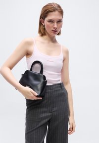 Femme aux cheveux courts roux portant un débardeur rose clair et un pantalon gris à fines rayures, tenant un petit sac à main noir sur un fond uni.
