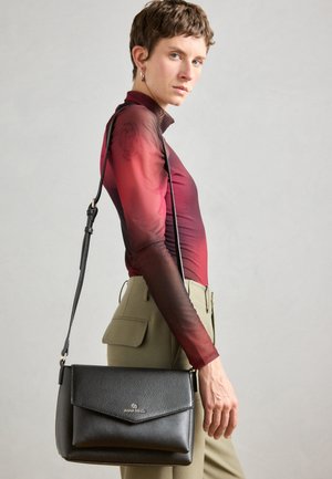 Black leather crossbody bag with a fold-over flap, textured surface, and gold logo. Model wears a gradient long-sleeve top and tailored pants.