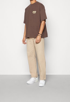 Ruskea yläosan t-paita, jossa edessä logo, yhdistetty beigeihin suoralahkeisiin housuihin ja valkoisiin tennareihin, minimalistinen muotoilu, rento istuvuus.