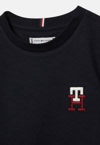 Černé tričko s krátkým rukávem a kulatým výstřihem s vyšitým logem "TH" v červené a bílé barvě na hrudi a štítkem Tommy Hilfiger uvnitř límce.