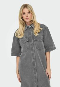 Grå denimklänning med korta ärmar, knäppning framtill och två bröstfickor. Har en avslappnad passform och klassisk kragsdesign.
