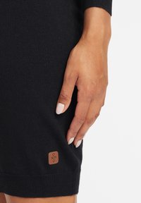 Abito maglione nero con una texture a coste. Una toppa con logo marrone è cucita sull'orlo. La mano poggia delicatamente sul tessuto.