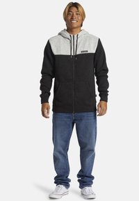 Haut à capuche zippé gris et noir avec poches avant, capuche avec cordons et un écusson de logo. Associé à un jean bleu et des baskets blanches.
