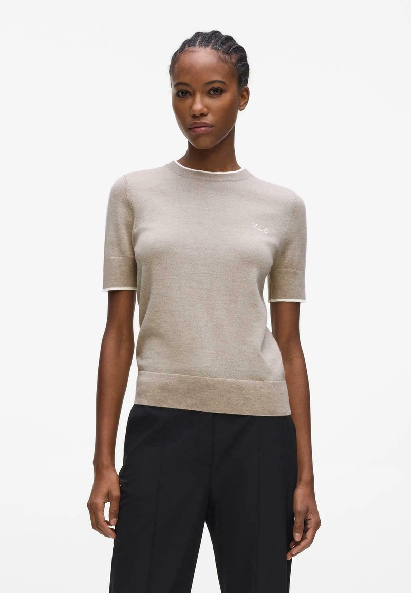 Femme portant un pull beige à manches courtes et un pantalon noir, debout avec les bras détendus sur un fond blanc.