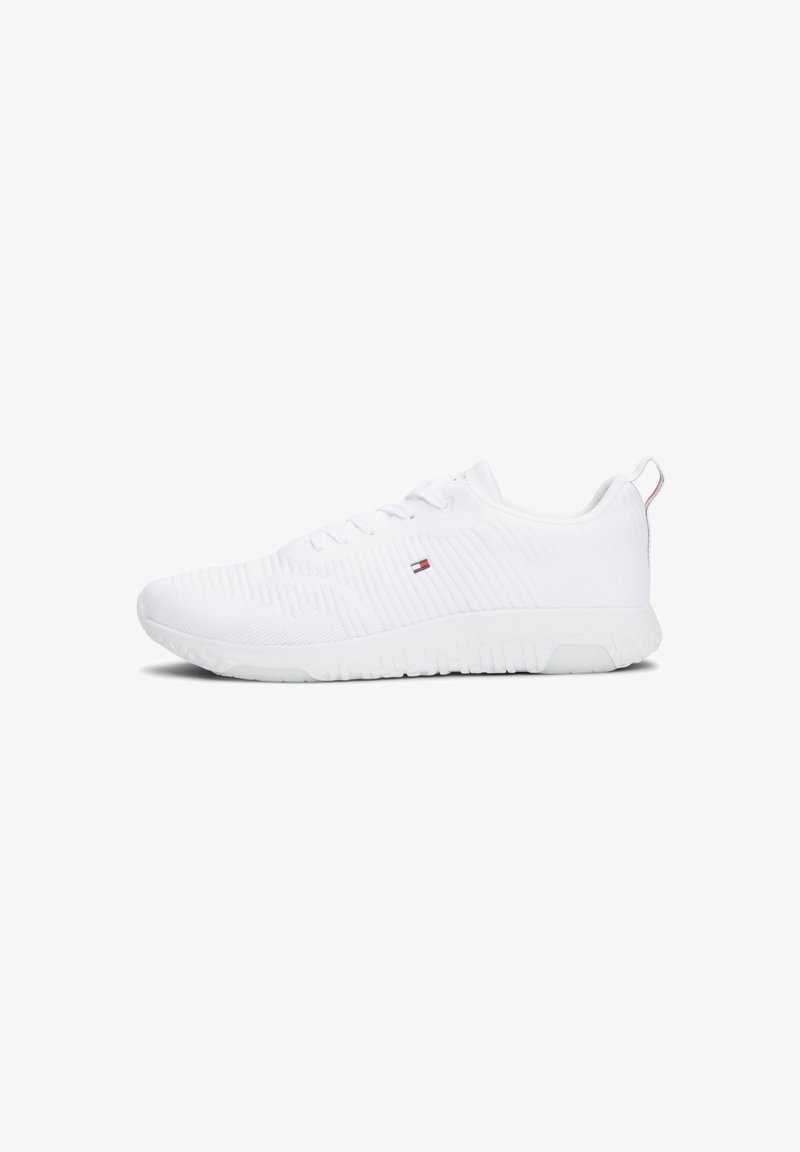Chaussure de sport blanche basse avec texture tricotée, lacets et petit logo rouge, blanc et bleu sur le côté, vue de profil.