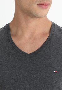 Camiseta de cuello en V de color gris oscuro en una tela suave. Presenta un pequeño logo bordado en colores rojo, blanco y azul en el lado izquierdo del pecho.
