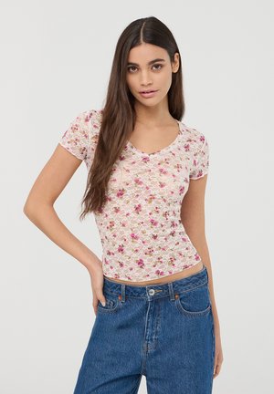 Top in pizzo con un motivo floreale rosa, maniche corte e scollo a V. Abbinato a jeans blu a vita alta. Texture morbida e traslucida.