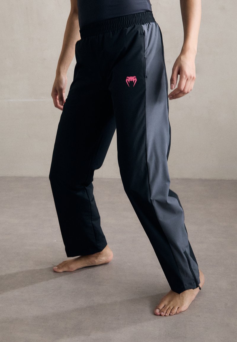 Sorte treningsbukser med en rosa logo, med elastisk midjebånd og tapered bein. Glatt, lett materiale og glidelåsdetaljer.