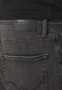 Tmavě šedé džínové kalhoty s černou koženou nášivkou označenou "JACK & JONES JEANS INTELLIGENCE," s jemným prošíváním a zadními kapsami.