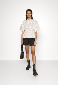 Blusa floral branca com cintura ajustada, combinada com calções pretos. A modelo segura uma mala preta e usa botas pretas chunky. Fundo simples.