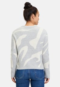 Grau- und cremefarbener gemusterter Pullover mit Rundhalsausschnitt, gerippter Textur und lässiger Passform, kombiniert mit blauen Jeans. Sichtbares rückseitiges Design.