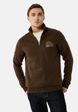 Joven con sudadera marrón con cremallera hasta el pecho con diseño de montaña y texto, combinado con vaqueros oscuros, de pie con las manos en los bolsillos.
