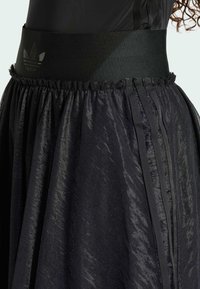 Black skirt featuring a textured, sheer fabric with pleats, an elastic waistband, and a subtle logo. Design emphasizes layered material.