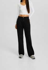 Jeans negros de pierna ancha hechos de mezclilla, con cintura alta y bolsillos delanteros, combinados con un top corto blanco de manga larga y zapatillas grises.