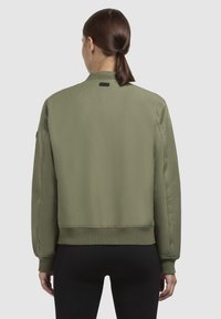 Olijf groene bomberjack met een gladde afwerking, ribbel manchetten en zoom, en een klein zwart label bij de kraag. Eenvoudig, minimalistisch ontwerp.