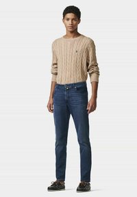 Pull en maille beige et jeans slim-fit bleu foncé. Le pull présente un motif en relief ; les jeans ont des poches avant et des détails de couture subtils.