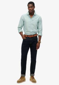 Camicia verde chiaro a maniche lunghe, jeans blu navy, cintura marrone e stivali beige. La camicia ha una tasca sul petto; i jeans sono aderenti e con i risvolti.
