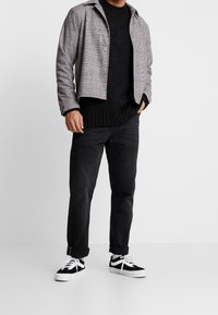 Svart stickad tröja tillsammans med en grårutig jacka, svarta denimjeans och svart-vita sneakers. Fokus på strukturerade tyger och mönster.