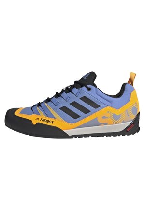 Chaussure de trail Adidas Terrex bleu et jaune avec semelle noire, trois bandes noires, fermeture à lacets à l'avant et tirant orange au talon.