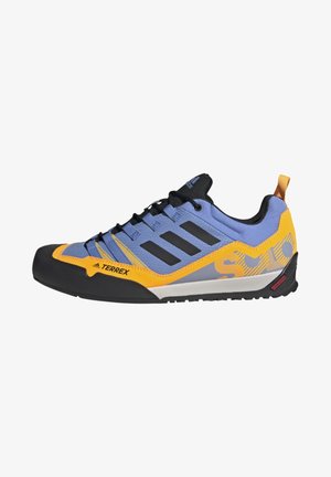 Chaussure de trail Adidas Terrex bleu et jaune avec semelle noire, trois bandes noires, fermeture à lacets à l'avant et tirant orange au talon.
