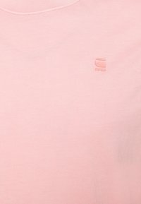 T-shirt rose clair en tissu doux, avec un col rond et un logo en relief discret en haut à gauche.