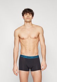 Svarte boxershorts med en turkis midjebånd som har "DIM"-logoen, laget av et glatt og mykt stoff. Modellen poserer foran en nøytral bakgrunn.