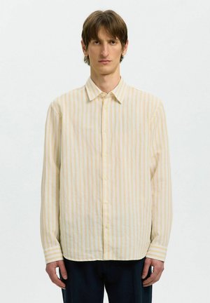 Homme debout devant un fond blanc, portant une chemise à manches longues à rayures jaunes et blanches avec des boutons, et un pantalon sombre.