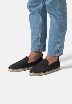 Personne portant des espadrilles noires slip-on avec une semelle tissée et un jean denim bleu clair coupé au-dessus des chevilles, debout sur un sol gris clair.