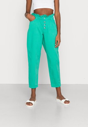Femme portant un pantalon taille haute turquoise avec des boutons et un top court blanc, debout avec une main dans la poche et des sandales blanches à enfiler.
