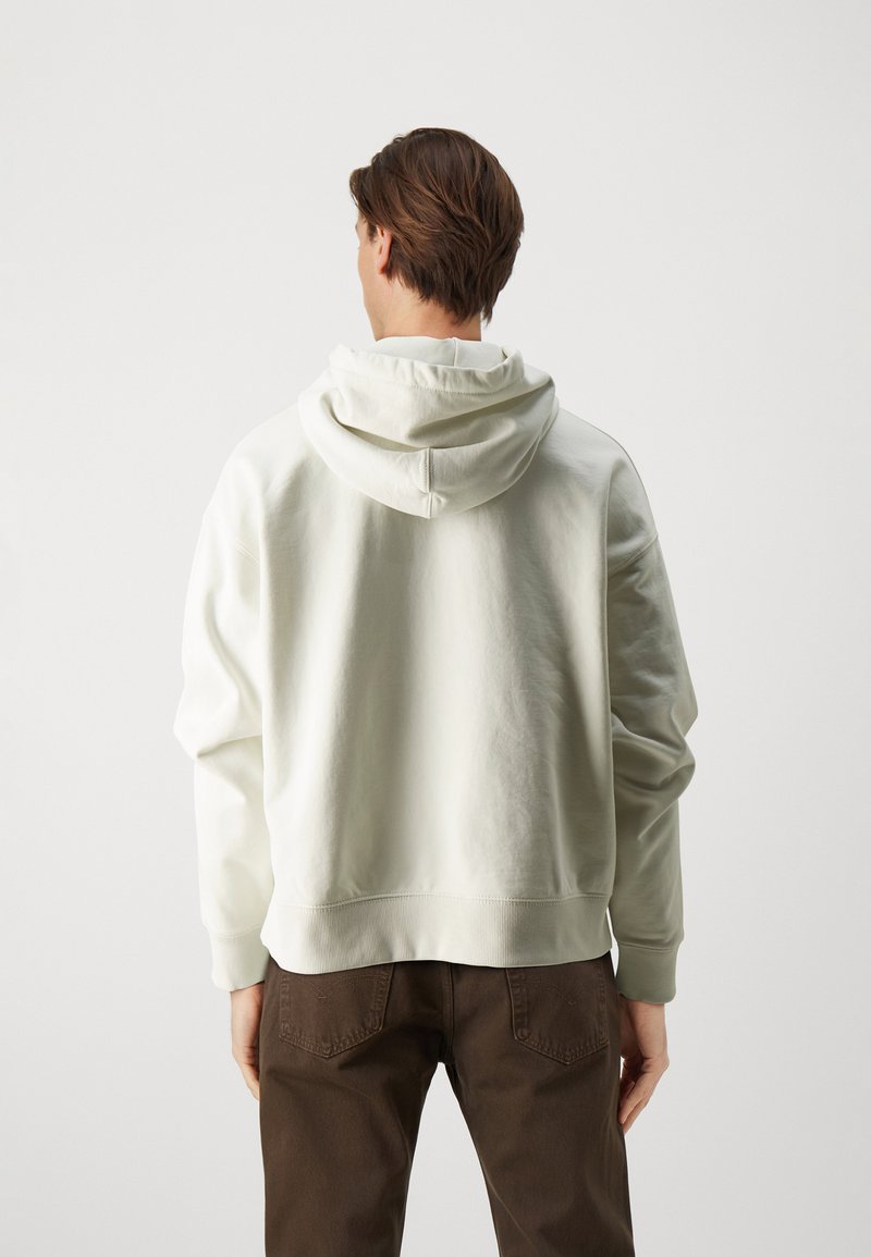 Crèmekleurige hoodie met een relaxte pasvorm, trekkoordcapuchon en geribde manchetten, gecombineerd met bruine broek. Soepele textuur, minimalistisch ontwerp.