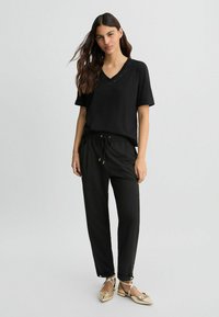 Schwarzes V-Ausschnitt-T-Shirt mit kurzen Ärmeln, kombiniert mit schwarzen Jogginghosen mit Kordelzug, die eine lockere Passform und eine glatte Textur aufweisen. Goldene flache Schuhe.
