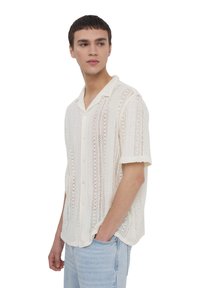 Maglietta a maniche corte di colore crema, realizzata in maglia con trama aperta texturizzata, colletto e chiusura con bottoni, abbinata a jeans azzurri chiari.
