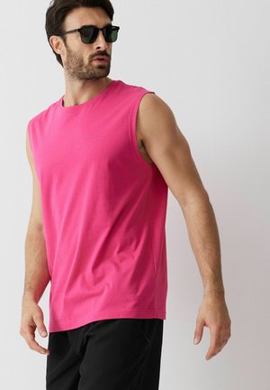 Mężczyzna w czarnych okularach przeciwsłonecznych, jaskrawo różowej koszulce bez rękawów i czarnych spodniach, stojący na jednolitym jasnym tle.