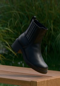 Must nahktest madala kontsaga pahkluu saabas, millel on soonilised detailid, ümar varvas ja madal plokkkonts. Riputatud puidust pinna kohale.