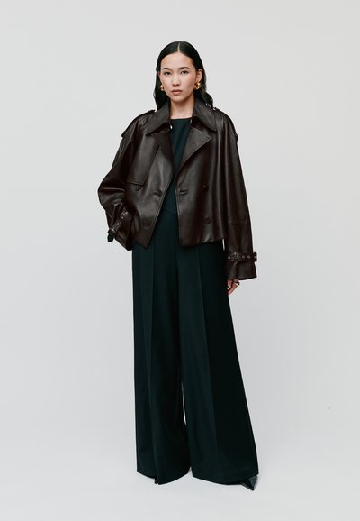 Dunkelbraune Lederjacke im Doppelreißverschluss-Stil mit Schulterklappen, kombiniert mit einem eng anliegenden schwarzen Overall mit weitem Bein und spitzen Schuhen.