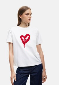 Witte katoenen T-shirt met een grote, rode, druipende hartafbeelding. Het shirt heeft een ronde halslijn en korte mouwen.