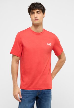 Mann trägt korallenrotes kurzärmeliges T-Shirt mit kleinem weißem Pferdelogo und blaue Jeans, steht mit einer Hand in der Hüfte vor weißem Hintergrund.