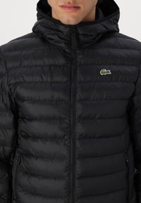 Juodas pūkinis švarkas su gaubtu, turintis siūtos tekstūros ir užtrauktuku užsegimą. Lacoste logotipas kairėje krūtinės pusėje. Šoninis kišenė su užtrauktuku.