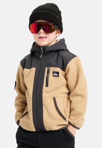 Chaqueta de forro polar para niños con cuerpo beige y hombros grises, con bolsillos con cremallera y capucha. Combinada con un gorro de punto negro y gafas de sol reflectantes.