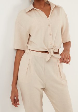 Femme portant une chemise beige en lin à manches courtes boutonnée, nouée à la taille, avec un pantalon plissé assorti taille haute.