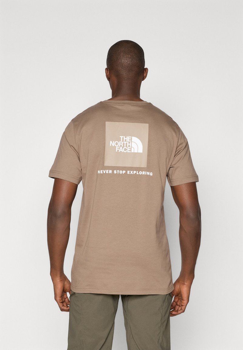 Tricou din bumbac maro cu mâneci scurte, având un logo dreptunghiular alb și textul "NEVER STOP EXPLORING" pe spate.