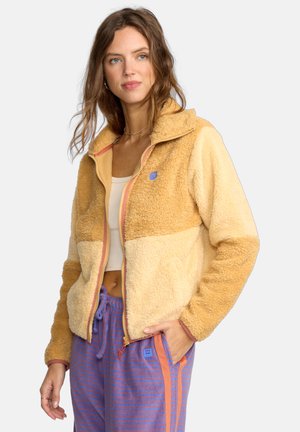 Fleece jacket in beige and light tan with a half-zip design, featuring a blue logo and contrasting pink zipper. Soft texture, relaxed fit.