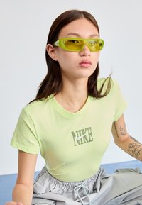Gai zaļš kokvilnas T-krekls ar teksturētu Nike logo priekšā, apvienots ar pelēkām biksēm ar auklu un dzeltenām saulesbrillēm.