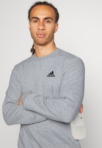 Šedá mikina s dlhým rukávom a okrúhlym výstrihom, s čiernym logom Adidas na hrudi. Tkanina vyzerá mäkko s mierne textúrovaným povrchom.