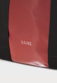 Sac noir avec un panneau rouge brillant, portant le mot "RAINS" en lettres argentées subtiles. Le matériau est lisse avec un design épuré.