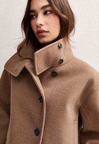 Manteau en laine marron avec un col haut et des boutons noirs, présentant une texture douce et un design minimaliste. Coutures accentuées visibles.