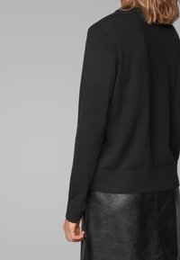Schwarzer Strickpullover mit Rundhalsausschnitt, gerippten Bündchen und Saum. Über einem eleganten, glänzenden schwarzen Rock getragen. Glatte Textur und klassisches Design.