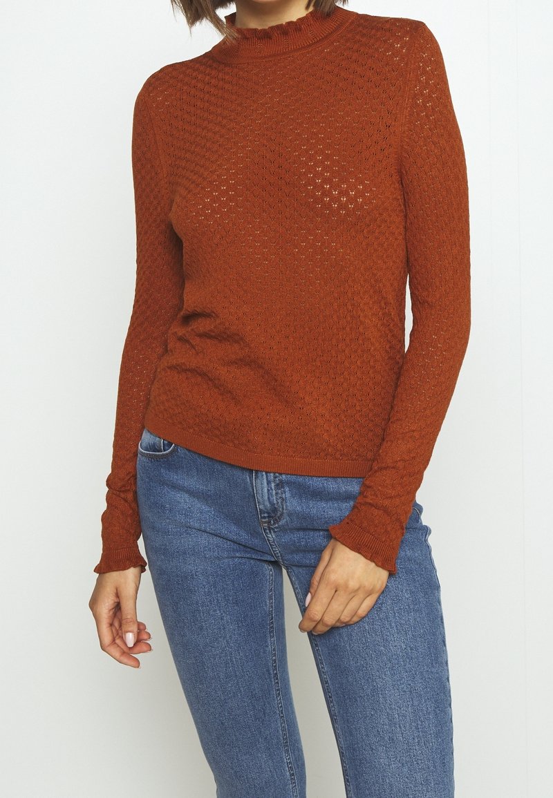 Pull en tricot couleur rouille avec un motif texturé, doté d'un col montant côtelé et de manches longues ajustées, associé à un jean bleu.